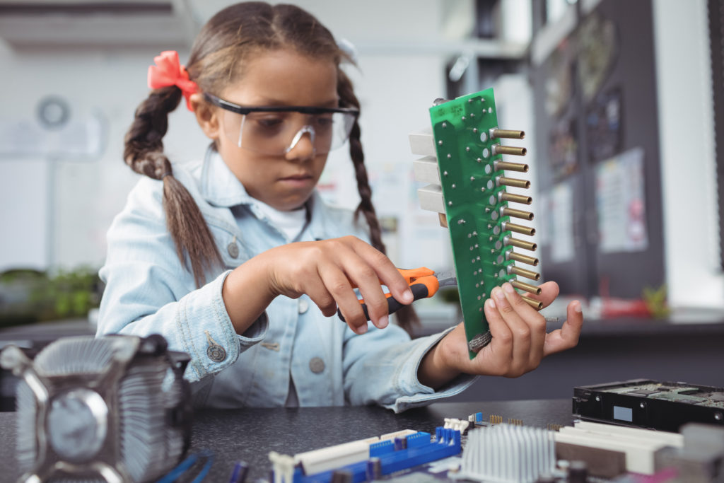 Concentrated elementary girl assembling circuit board on desk at electronics lab